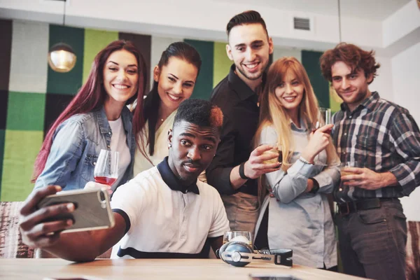 Přátelé se baví v restauraci. Tři kluci a tři holky dělají selfie a smějou se. V popředí chlapec drží chytrý telefon. Všichni noste ležérní oblečení Stock Snímky