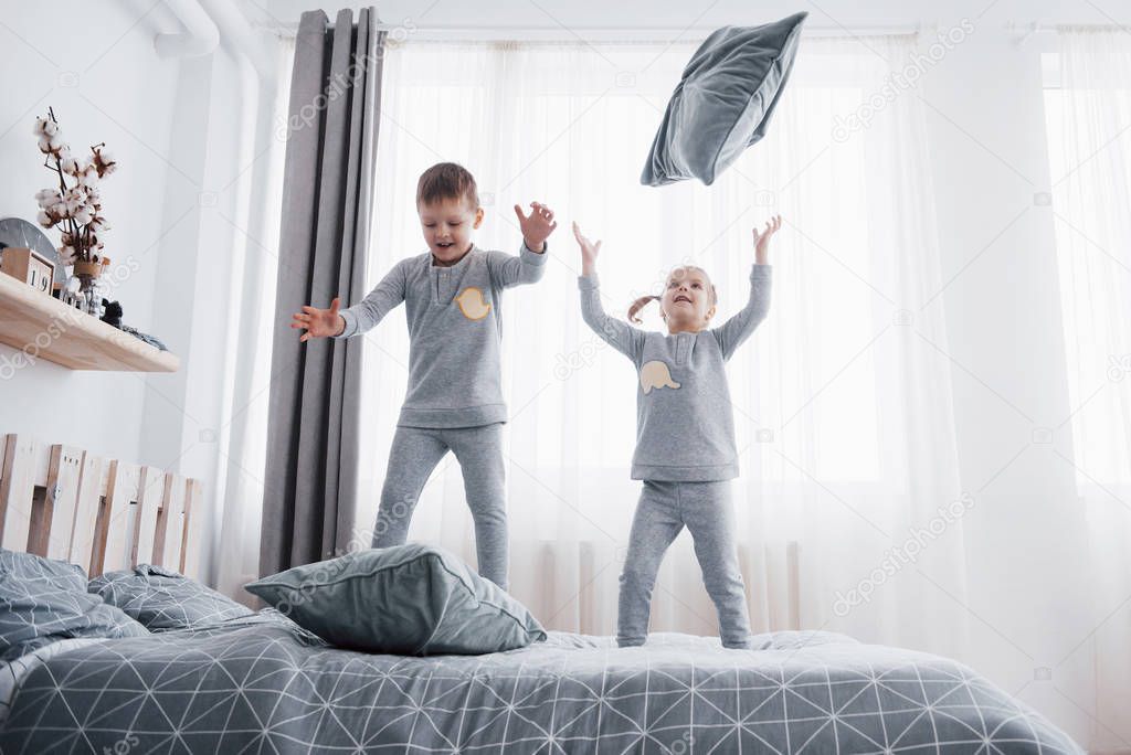 Happy kids playing in white bedroom. Little boy and girl, brother and sister play on the bed wearing pajamas. Nightwear and bedding for baby and toddler. Family at home.