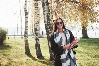 Çalışma ve dinlenme kavramı. Güneş gözlüklü genç esmer ağaçların yanındaki parkta duruyor ve not defteri tutuyor..