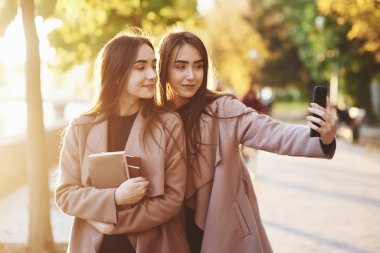 Genç, gülümseyen esmer ikiz kızlar siyah bir telefonla selfie çekerken, bir tanesi elinde kitap destesi tutuyor, gündelik palto giyiyor ve bulanık arka plandaki sonbahar güneşli park sokağında dikiliyor..