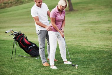 Şimdi kendin dene. Erkek kadına spor sahasında arkada ekipmanla golf oynamayı öğretiyor..