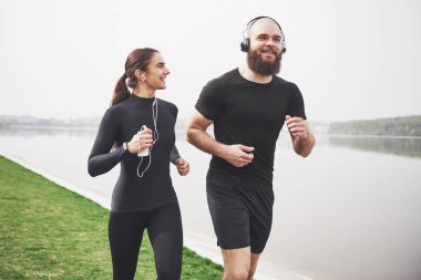 Profile view image of an athletic couple wearing arm bands with smart phones and headphones. Young bearded man and woman exercising together in morning.