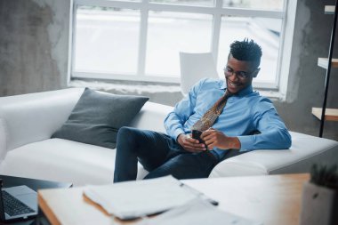 Close up. Smiling afroamerican guy sitting on the couch and looking at his smartphone.