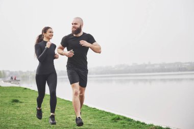 Couple jogging and running outdoors in park near the water. Young bearded man and woman exercising together in morning.