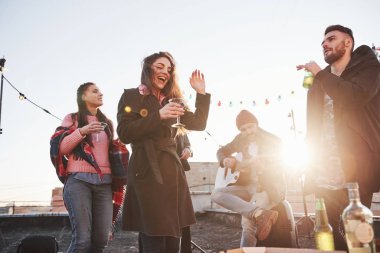 Dans ve şarkı. Alkol içen ve gitar çalan bir çatı partisi. Arkasında güneş ışığı.