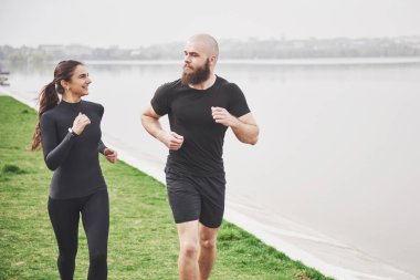 Couple jogging and running outdoors in park near the water. Young bearded man and woman exercising together in morning.