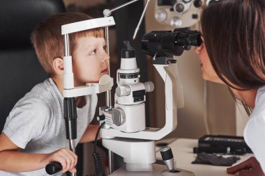 This might take one more minute. Little boy have test for his eyes with special optical apparatus by female doctor.