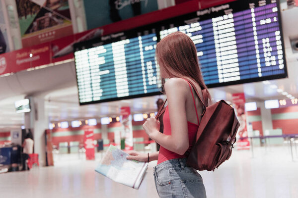 Young woman in airport with map in hands and information board on the background is looking to the side.