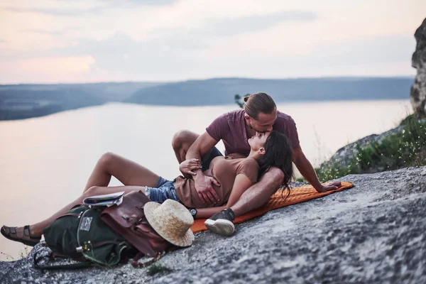 キスのために横になって若いカップルは 湖を背景に豪華な岩の端に積極的に休暇を過ごすことにしました — ストック写真