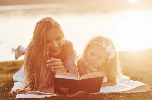 Stumbled upon a joke. Mom and daughter reading a book in a very sunny day laying on the grass with lake at background.
