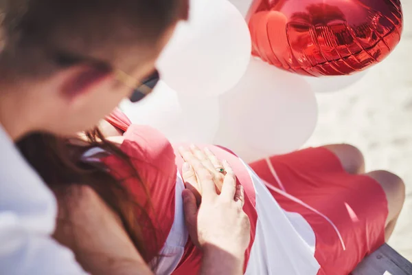 赤ちゃんを待ってる 最愛の夫を持つ妊婦がベンチに座る 夫の手は丸い腹の妻を包含する 赤い弓で桜の白いドレスを着た妊婦さん 接近中だ 9ヶ月 — ストック写真