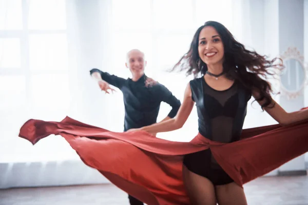 Girl is spin around herself. Young pretty woman in red and black clothes dancing with bald guy in the white room.