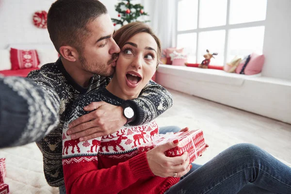 プレゼントを持ちながら彼氏のところに歩いている女性 男は彼と彼の妻のクリスマスの服を着て 装飾的な美しい部屋の床に座って自撮り — ストック写真