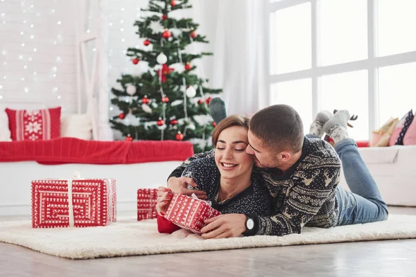 妻にクリスマスプレゼントを素敵な若いカップルが住んでいる部屋に横たわっています緑の休日の木を背景に — ストック写真