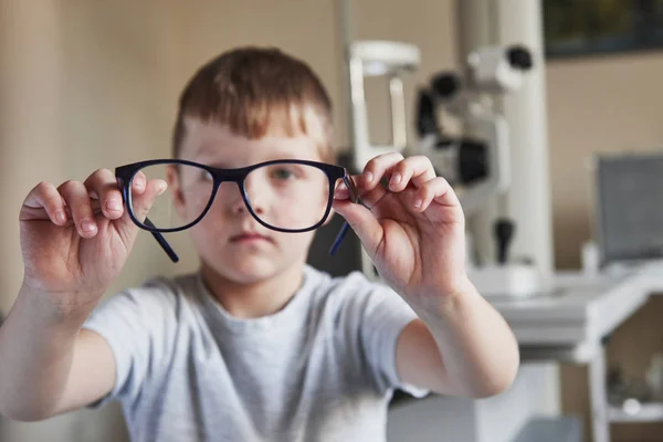 男の子は背景に眼科機器と彼の細長い手に眼鏡を保持しています — ストック写真