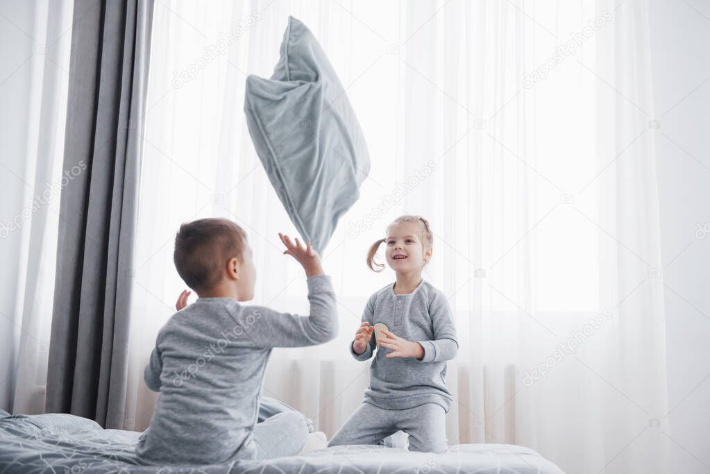 Happy kids playing in white bedroom. Little boy and girl, brother and sister play on the bed wearing pajamas. Nursery interior for children. Nightwear and bedding for baby and toddler. Family at home