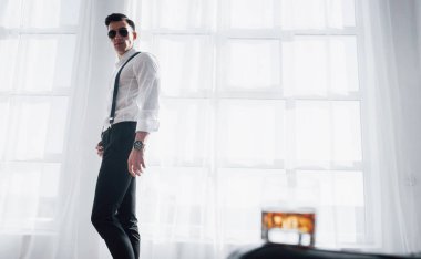 Doğruca kameraya. Odadaki şık saç kesimli adam pencerenin yanında duruyor. Önünde içki olan bir bardak..