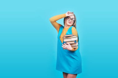 Kafası karışmış, gözlüklü şirin bir kız elinde renkli mavi arka planda izole bir kitap yığını..