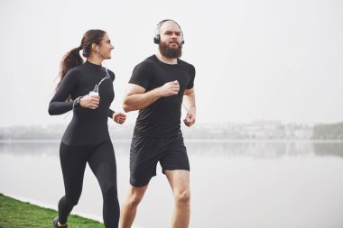Couple jogging and running outdoors in park near the water. Young bearded man and woman exercising together in morning.