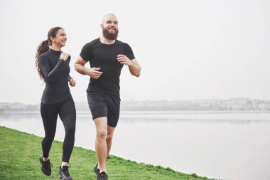 Couple jogging and running outdoors in park near the water. Young bearded man and woman exercising together in morning.