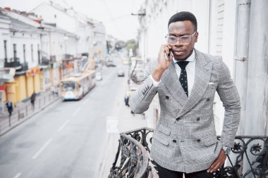 Resmi giyinmiş genç Afrikalı adam cep telefonuyla konuşuyor ve dışarıda dururken gülümsüyor..