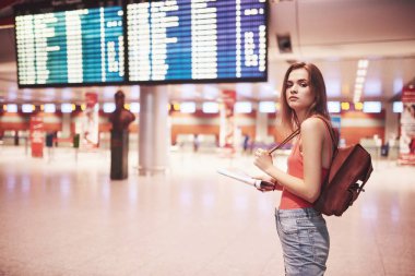 Uluslararası havaalanında sırt çantalı güzel bir genç turist kız, uçuş bilgi panosunun yanında..
