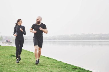 Couple jogging and running outdoors in park near the water. Young bearded man and woman exercising together in morning.