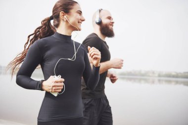 Profile view image of an athletic couple wearing arm bands with smart phones and headphones. Young bearded man and woman exercising together in morning.