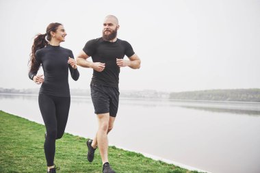 Couple jogging and running outdoors in park near the water. Young bearded man and woman exercising together in morning.