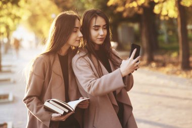 Genç, güzel, esmer ikiz kızlar birbirlerine bakıyor ve siyah telefonlarıyla selfie çekiyorlar. Bu arada içlerinden biri elinde kitap tutuyor, palto giyiyor ve bulanık arka plandaki sonbahar güneşli park yolunda dikiliyor..