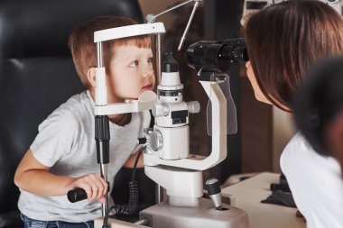 Please don't move your eyes. Little boy have test for his eyes with special optical apparatus by female doctor.
