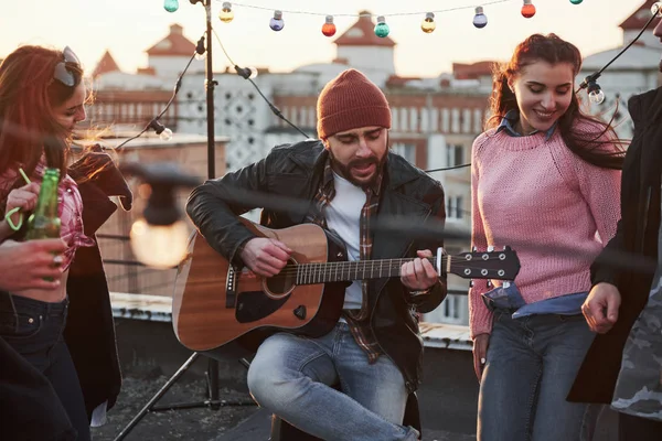 Gitarist çatıdaki arkadaşları için dekoratif renkli ampullerle oturup şarkı söylüyor..