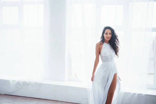 Looking straight into the camera. Beautiful woman in white dress stands in white room with daylight through the windows.