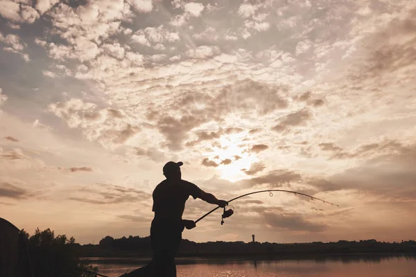 Silhouette of fisherman throws a fishing pole into the lake at sunset