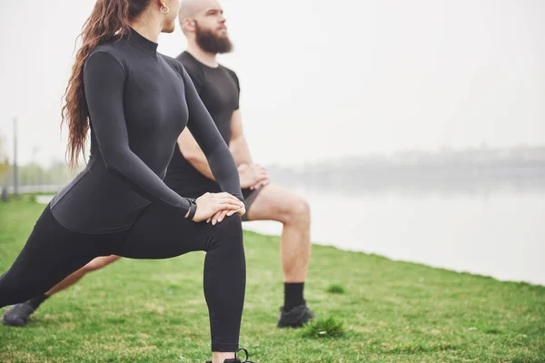 Fitness couple stretching outdoors in park near the water. Young bearded man and woman exercising together in morning.