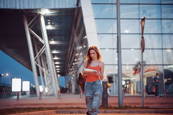 Young girl costs at night near the terminal of the airport or station and reading city map and looking for hotel. Cute tourist with backpacks determine the concept of travel.