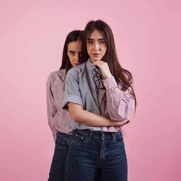 Stands in the studio with pink background. Second girl behind her sister.