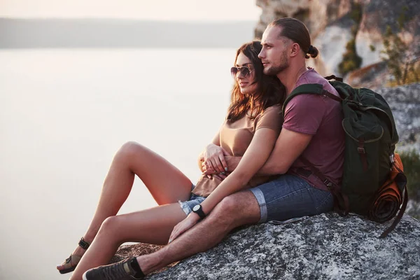 Hugging couple with backpack sitting on top of rock mountain enjoying view coast a river or lake. Traveling mountains and coast, freedom and active lifestyle concept.