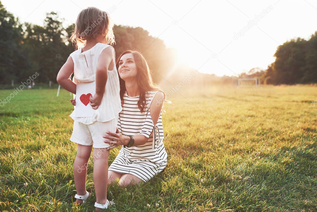 Beautiful sunlight above the trees. Girl hides the book with red heart on the back from the smiling sitting mother.