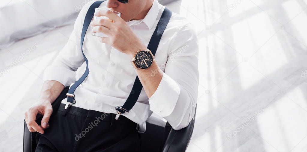 Cropped photo of drinking beverage. Young stylish businessman sitting on a black chair with glass of whiskey.