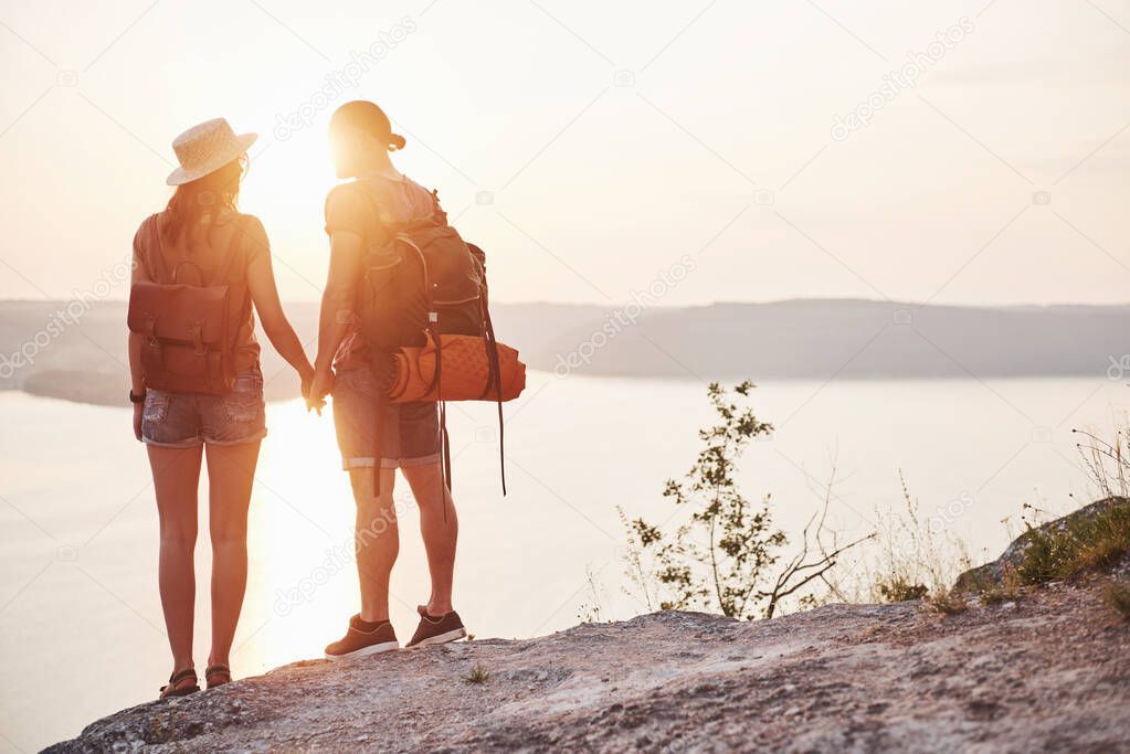 Two tourist male and woman with backpacks stand to the top of the crag and enjoying sunrise. Traveling mountains and coast, freedom and active lifestyle concept.