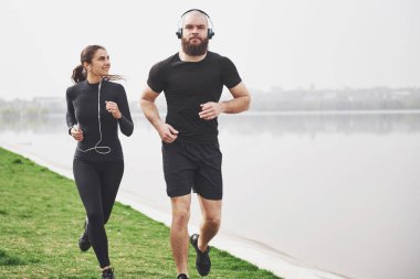 Couple jogging and running outdoors in park near the water. Young bearded man and woman exercising together in morning.