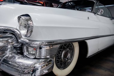 Fotoğraf sol taraftan. Otomobil fuarında kapalı park edilmiş beyaz antika bir arabanın önünde..