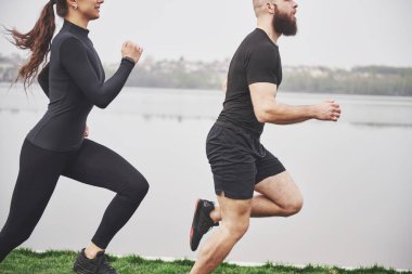 Couple jogging and running outdoors in park near the water. Young bearded man and woman exercising together in morning.