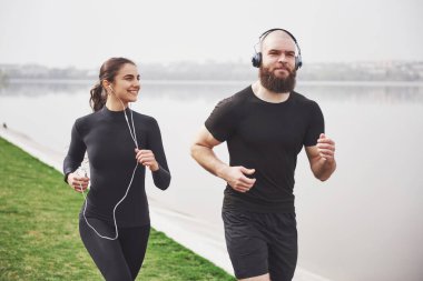 Profile view image of an athletic couple wearing arm bands with smart phones and headphones. Young bearded man and woman exercising together in morning.