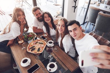 Bir grup insan bir kafede selfie çekiyor. En iyi arkadaşlar yemek masasında toplanmış pizza yiyor ve çeşitli içkiler söylüyorlardı..