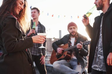 Güzel mutlu kıza odaklan. Alkol içen ve gitar çalan bir çatı partisi. Arkasında güneş ışığı.