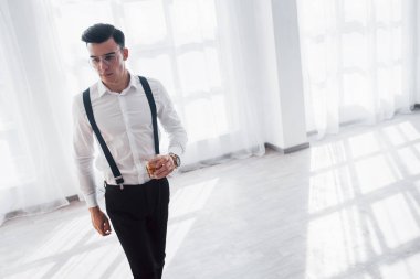 Boş oda. Lüks takım elbiseli bir adam elinde viskiyle beyaz bir odada yürüyor..