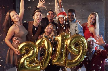 Yeni Yıl yaklaşıyor! Noel Baba şapkalı, altın renkli sayılar taşıyan ve partiye konfeti atan neşeli, etnik çeşitliliğe sahip bir grup genç..