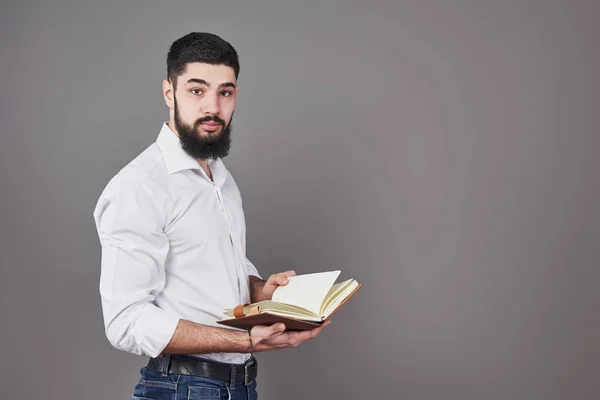 Portrait of a bearded young man wearing a white shirt and holding an open planner and a pen. A gray wall background.